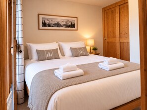 Una cama bien hecha con ropa blanca y dos toallas dobladas, una lámpara de noche, un cuadro con montañas y un armario de madera.