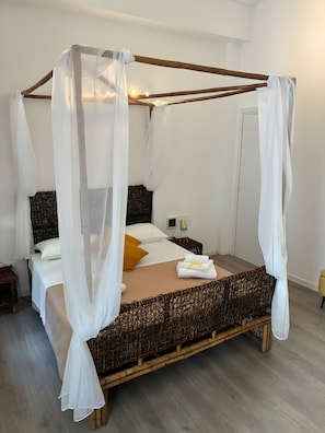 Ein Bett mit Baldachin, drapiert mit weißen Stoffbahnen, ein hölzernes Kopfteil, ein Nachttisch mit Lampe und ein kleiner Korbwannenkorb.