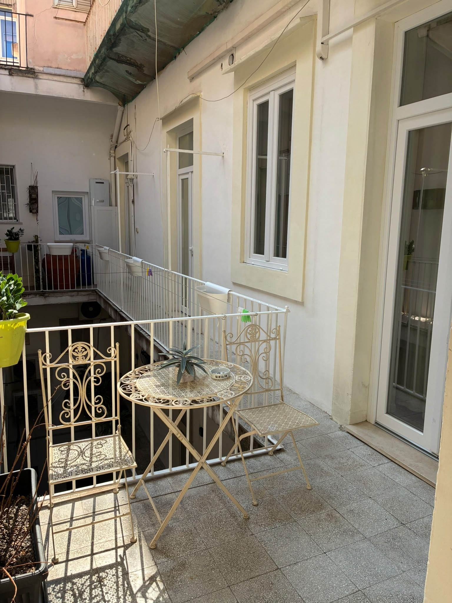A balcony with a metal railing, a round table, and chairs, a potted plant, and a window with a view of the street.
