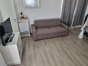 A room with a sofa, a small table with a cup and saucer, a window with curtains, and a desk with a computer and remote controls. - Room 'Camera Dea' with Balcony, Wi-Fi and Air Conditioning (Siracusa)