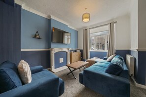 Ein Zimmer mit blauer und weißer Farbgestaltung, ausgestattet mit einer Couch, einem kleinen Tisch, einem Fernseher und einem Heizkörper.