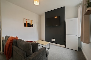 Ein kompakter Wohnbereich mit einer Couch, einem kleinen Tisch, einem flachbildfernseher an der Wand und einem weißen Kühlschrank.