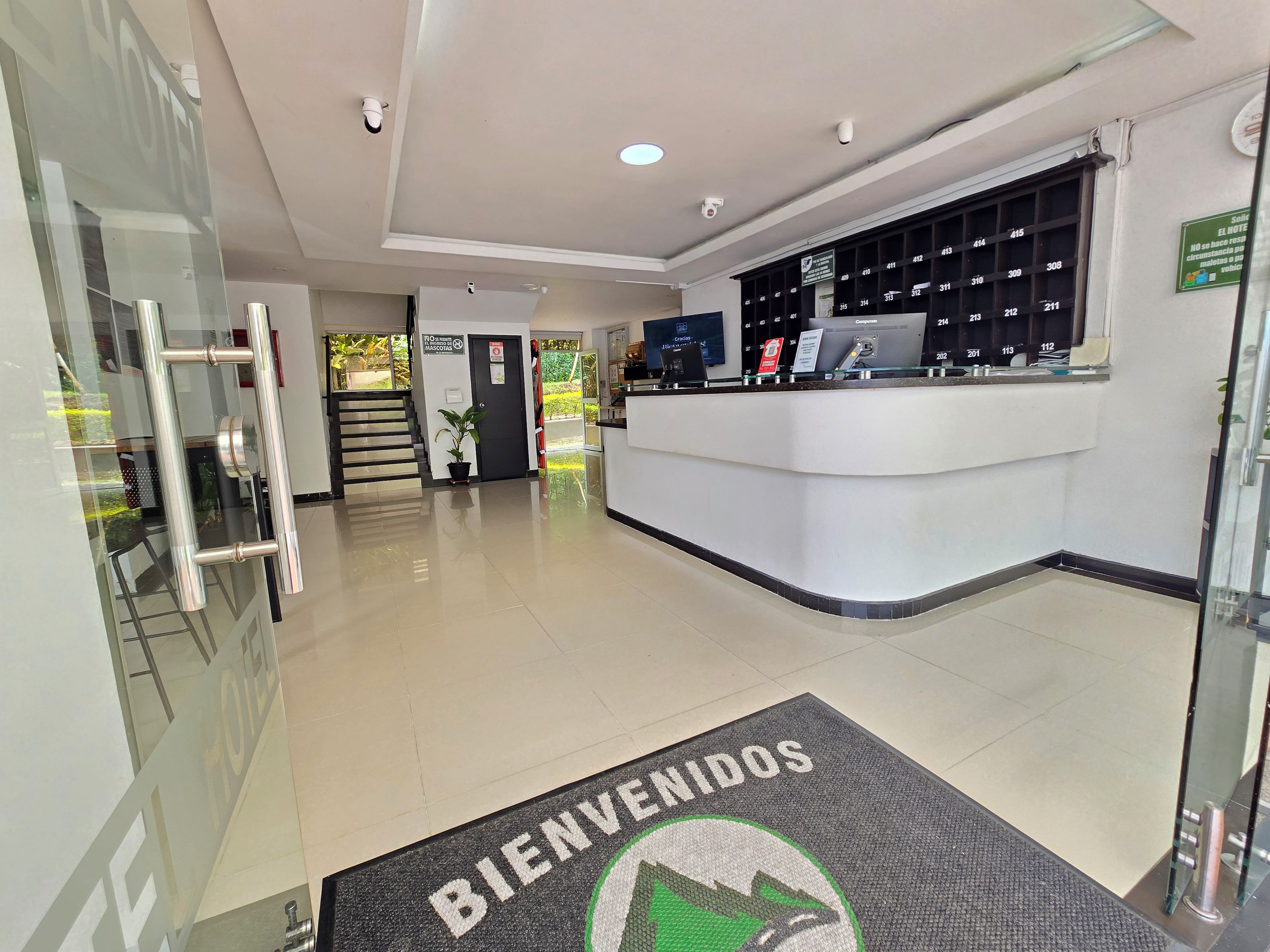 A hotel reception area with a reception desk, a 'BIENVENIDOS' mat, a staircase, and a glass door.