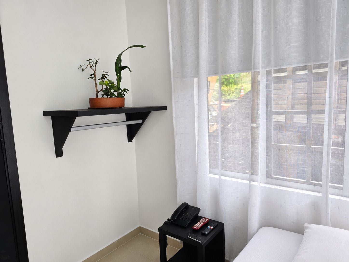 Ein Hotelzimmer mit einem Bett, einem kleinen Nachttisch mit Telefon und Fernbedienungen, einem Fenster mit durchsichtigen Vorhängen und einem an der Wand befestigten Regal mit einer Topfpflanze.