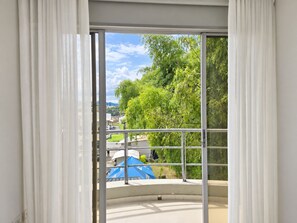 A view through a window with white curtains, revealing a balcony with a blue umbrella and greenery. - Denaka Hotel (Dosquebradas)