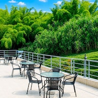 Un patio al aire libre con mesas y sillas redondas, rodeado de abundante vegetación y una barandilla metálica.