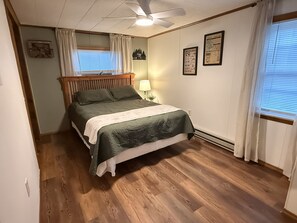 Quarto com cama, mesa de cabeceira, ventoinha no teto e janela com cortinas.
