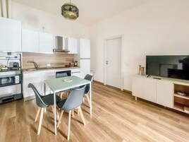 Una cocina moderna con armarios blancos, una mesa de comedor con sillas, horno empotrado y un televisor de pantalla plana sobre un mueble.