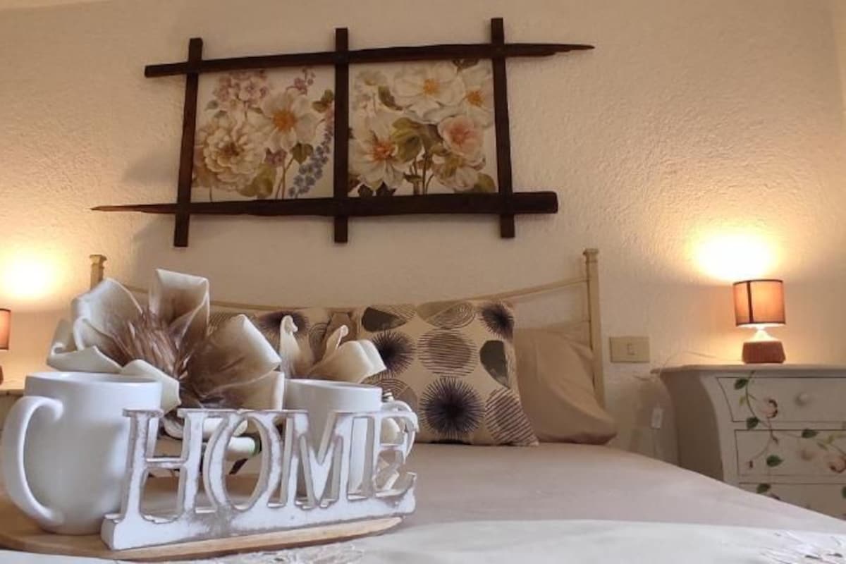 A neatly made bed with a 'HOME' sign, two bedside tables with lamps, and a floral artwork on the wall.