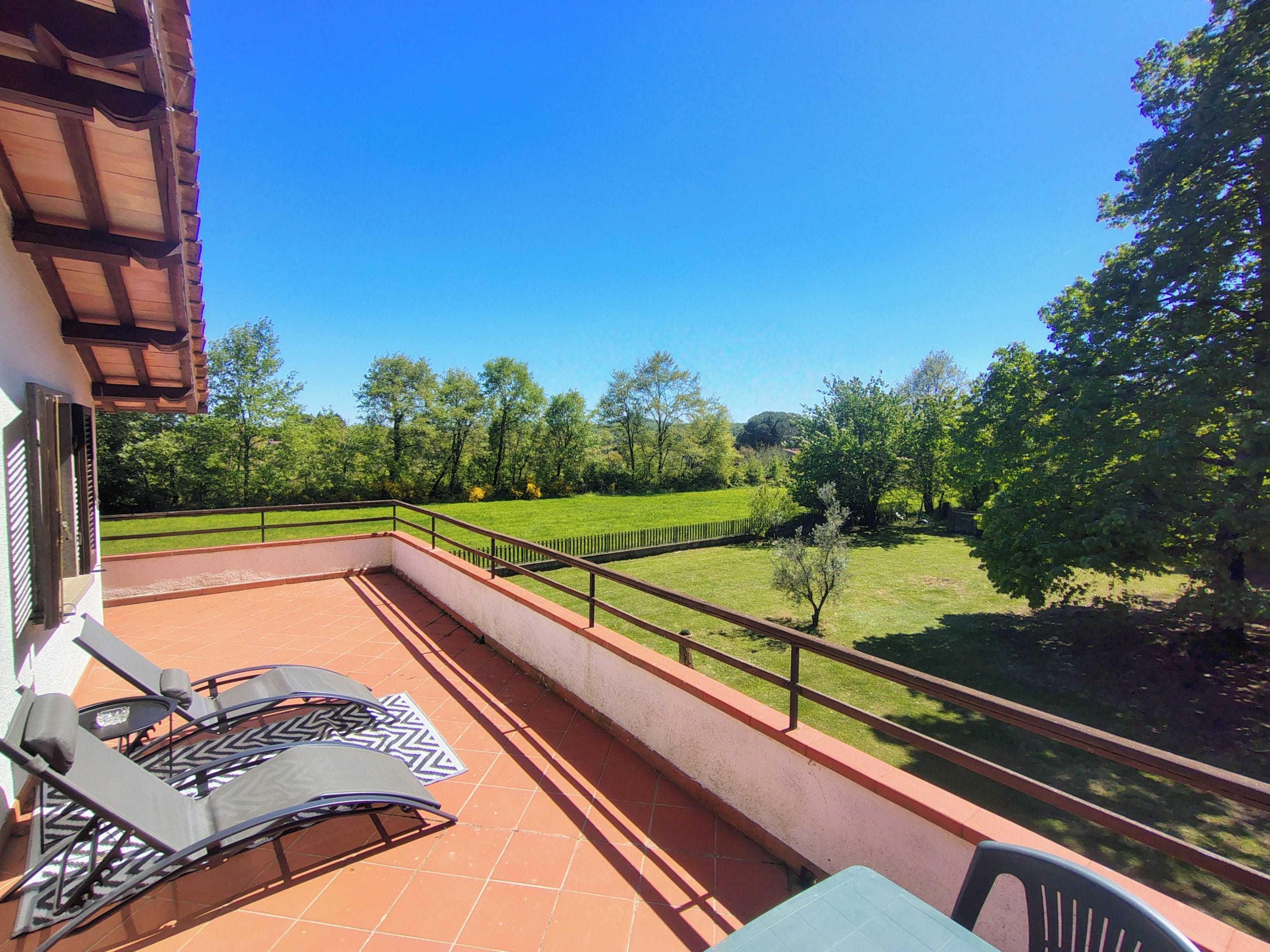 Un balcone con vista su un prato verde e alberi, un tavolo con sedie e una ringhiera.