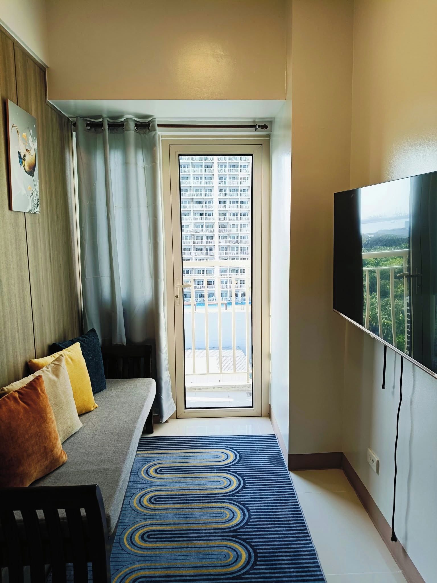 A room with a sofa, a television, a patterned rug, and a view of a building through a door.