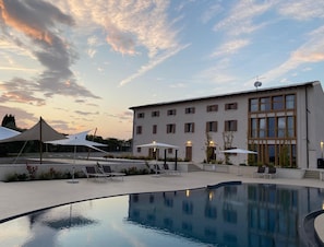 Zona de piscina con sillas de descanso y sombrillas, un edificio con múltiples ventanas y cielo despejado.