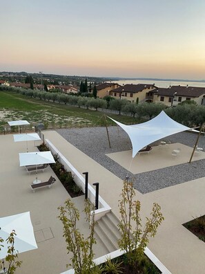 Una terraza exterior con múltiples tumbonas y sombrillas, un dosel blanco y vistas al mar y edificios al fondo.