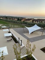 Μια υπαίθρια βεράντα με πολλές ξαπλώστρες και ομπρέλες, μια λευκή τέντα και θέα στη θάλασσα και στα κτίρια που βρίσκονται στο βάθος.