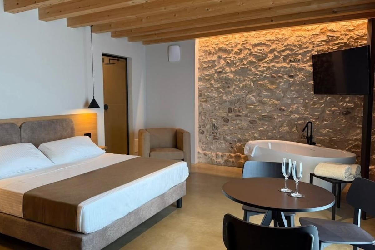 A bedroom with a bed, a chair, a round table with glasses, and a stone wall.