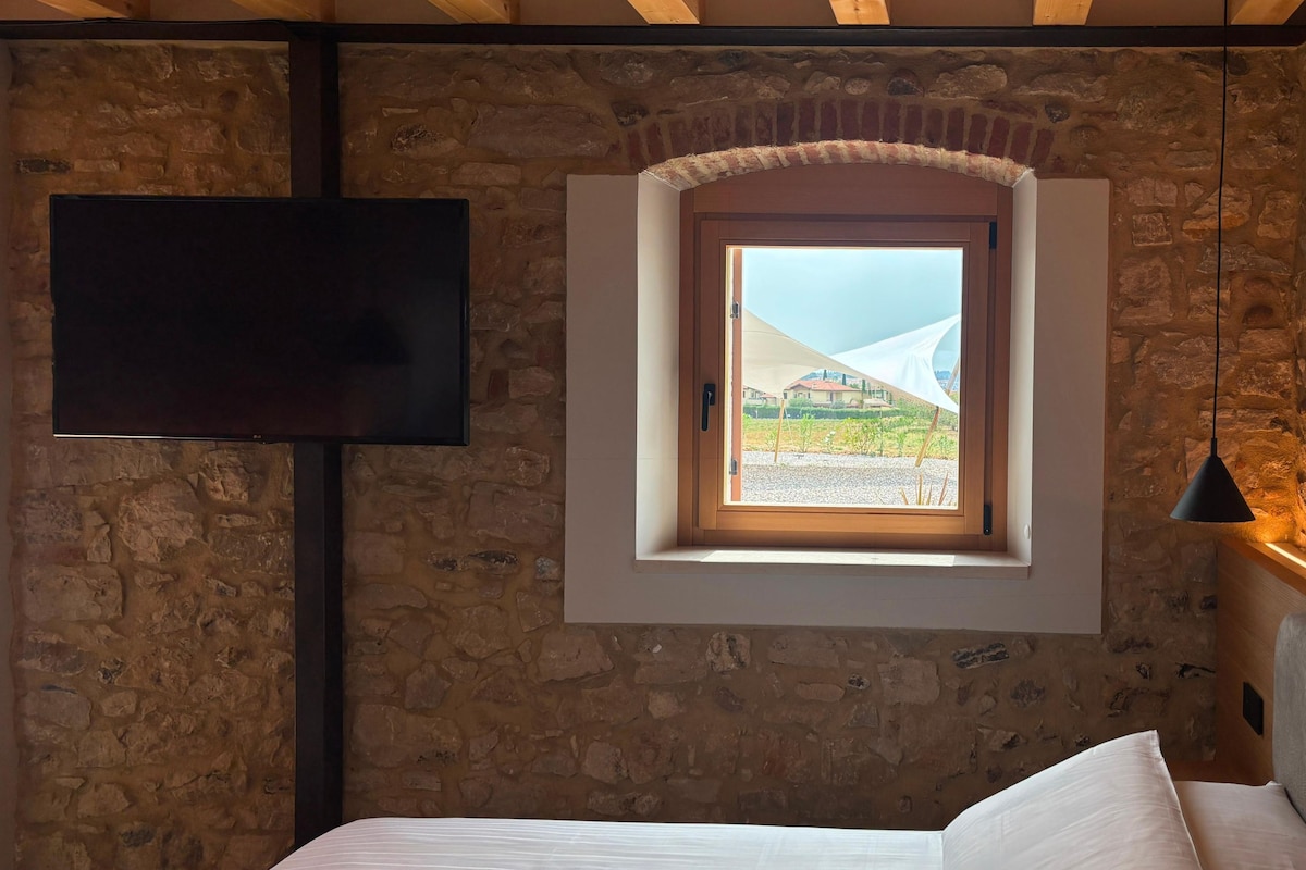 A bedroom with a stone wall, a TV mounted on the wall, a window with a view of buildings, and a bed with white bedding.