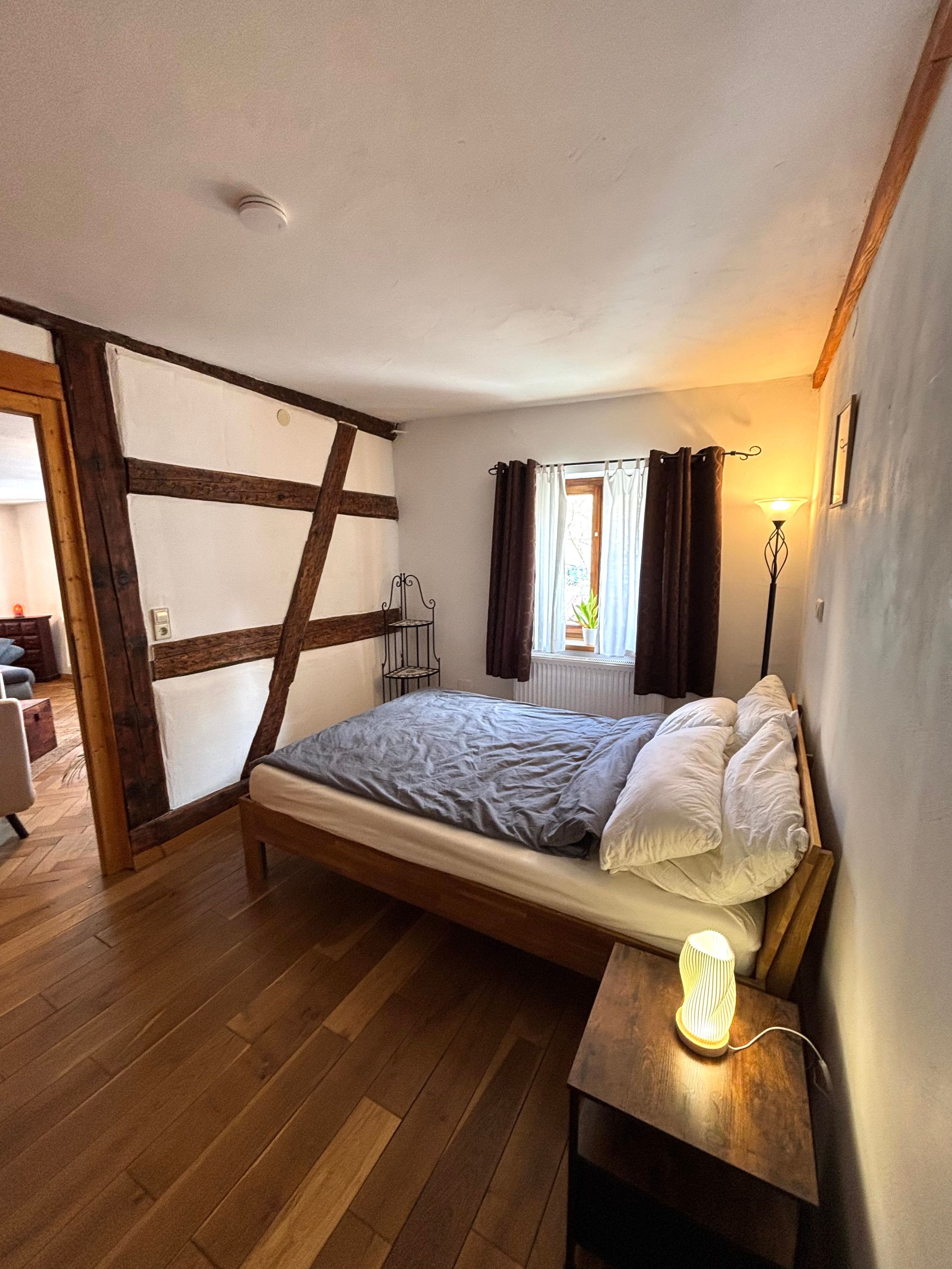 Un dormitorio con cama de madera, una mesita de noche con lámpara, una ventana con cortinas y una puerta que da a otra habitación.