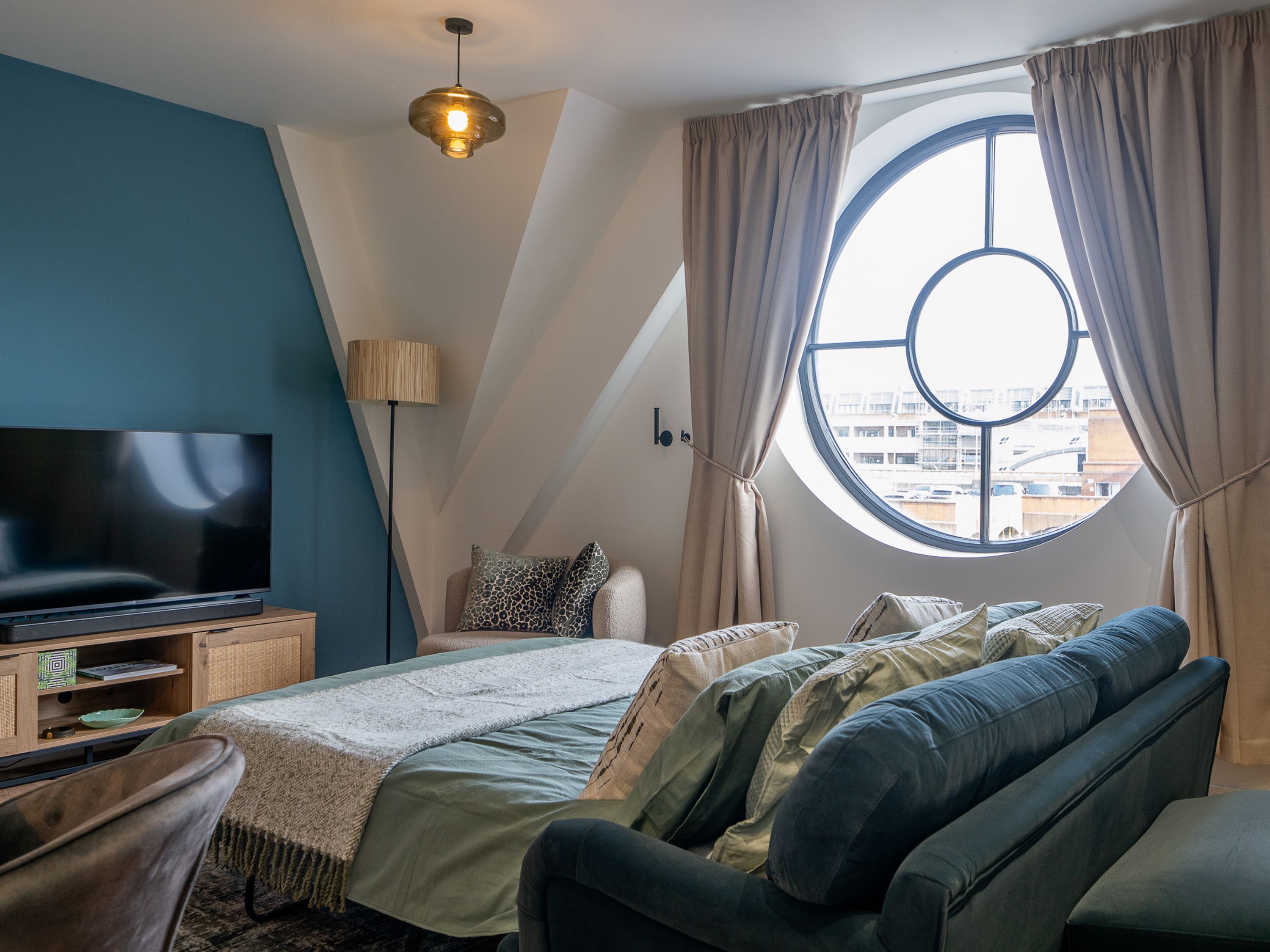 Ein modernes Schlafzimmer mit einem großen Bett, einem Flachbildfernseher, einem Fenster mit Vorhängen und Blick auf Gebäude.