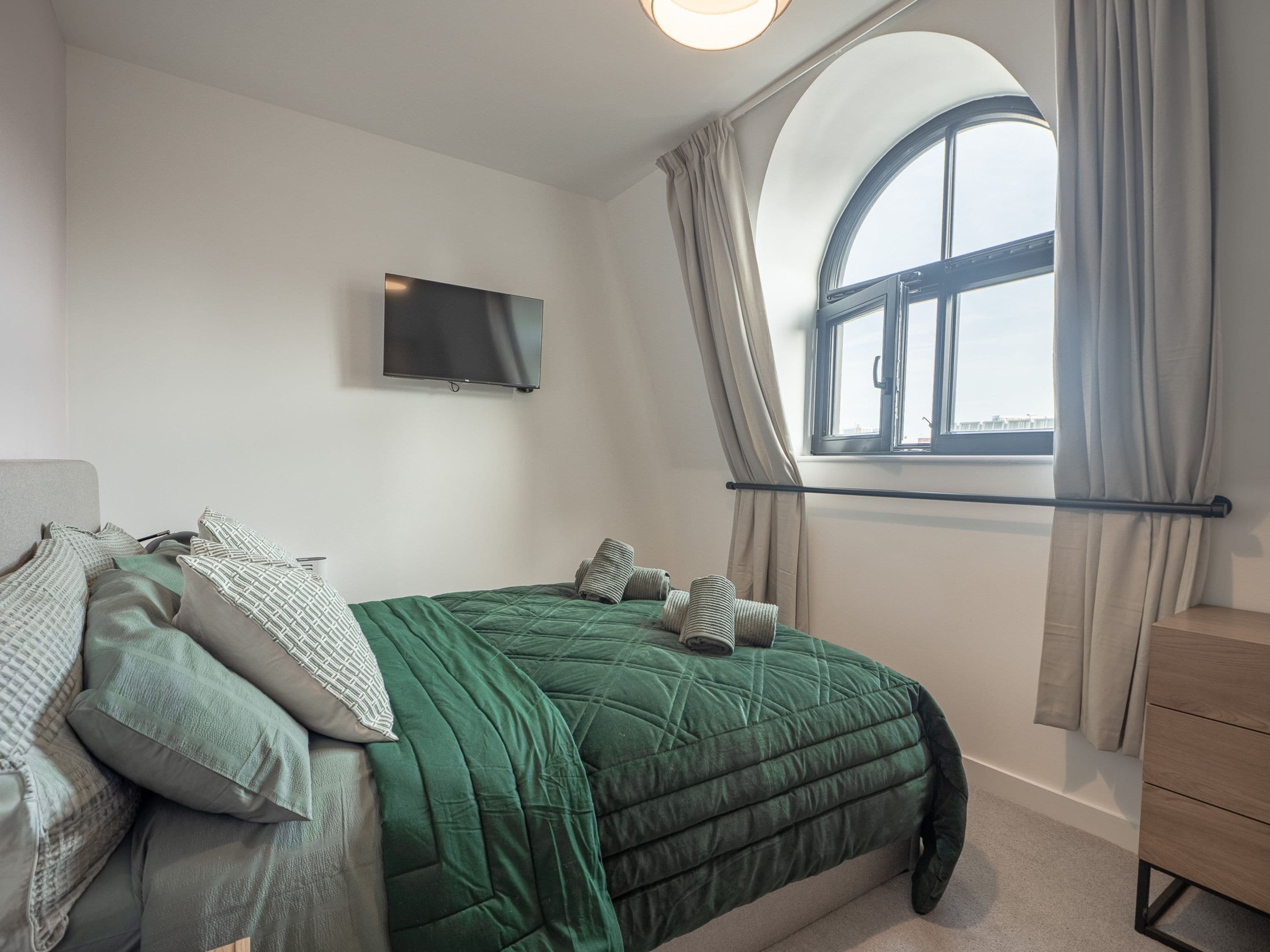 Ein Schlafzimmer mit einem Bett, einem Fernseher, einem Fenster mit Vorhängen und einer Kommode.
