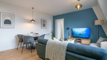 Ein modernes Wohnzimmer mit blauer Akzentwand, einem Fernseher, einem Essbereich mit Stühlen und einem Sofa.