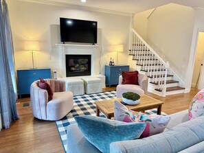 A living room with a fireplace, a flat-screen TV, a staircase, and a sofa with cushions. - Come experience this spacious 3-bedroom, 3-bath home in the heart of Starkville, where comfort, style, and a prime location create the perfect stay. Just 5 minutes to MSU, Downtown Starkville & The Cotton District, with plenty of parking. (Starkville)