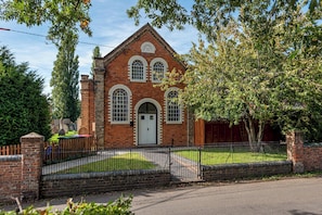 A brick chapel with a white door and arched windows, surrounded by a metal fence and trees. - The Old Chapel | A Stay Like No Other - Pass the Keys (Edgmond)