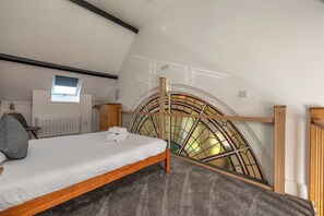Une chambre avec un plafond en pente, un lit, un radiateur, une petite fenêtre et une fenêtre à vitraux.