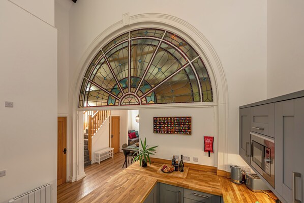 Una cocina amplia con encimera de madera, un comedor con mesa y sillas, y un gran ventanal de arco con vidrieras.