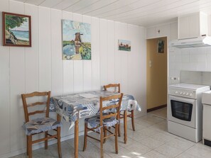 A dining area with a table set for four, wooden chairs, and a white tiled kitchen with a stove. - Vrbo Property (Beaussais-sur-Mer)