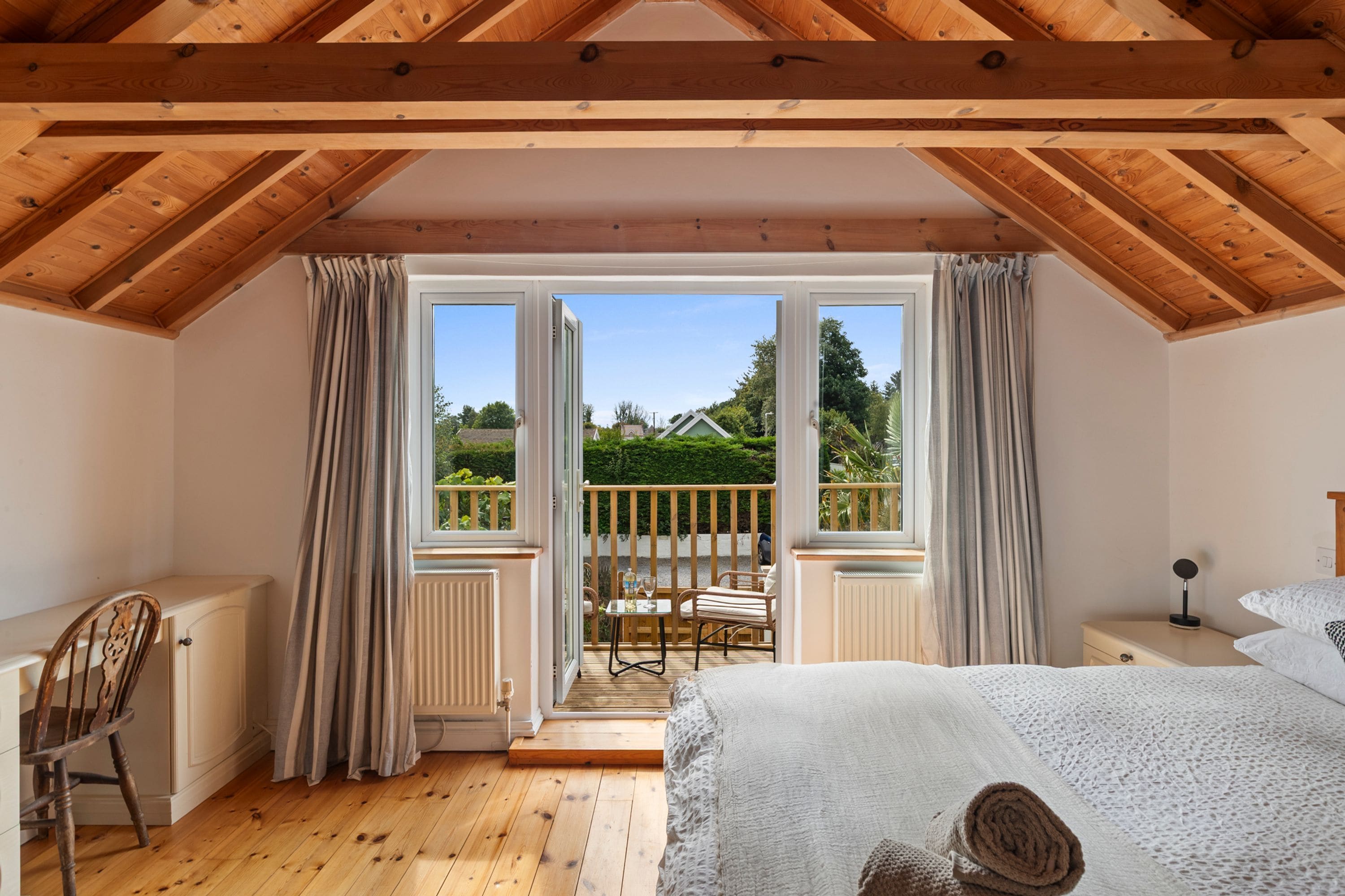 Quarto com teto de madeira, uma cama, uma cadeira, uma mesa com cadeiras e vista para o exterior por uma grande janela.