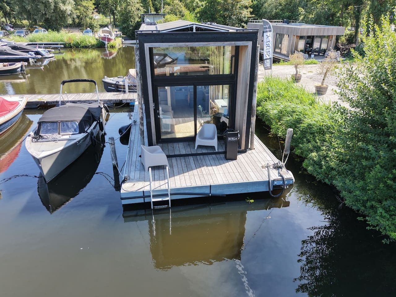 A floating house with a glass enclosure, docked near a calm water body, surrounded by greenery.