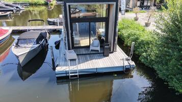 A floating house with a glass enclosure, docked near a calm water body, surrounded by greenery.