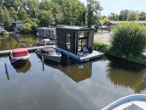 Een drijvend woonschip afgemeerd aan een rustig waterlichaam, omringd door groen en een heldere lucht.