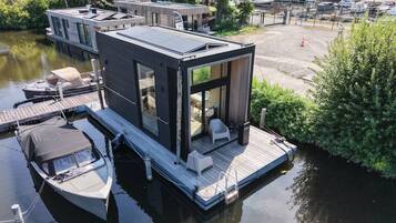 Una casa flotante moderna con terraza, una pequeña embarcación amarrada cerca y una vista despejada del agua y la vegetación circundante.