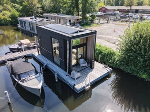 Una casa flotante moderna con terraza, una pequeña embarcación amarrada cerca y una vista despejada del agua y la vegetación circundante.