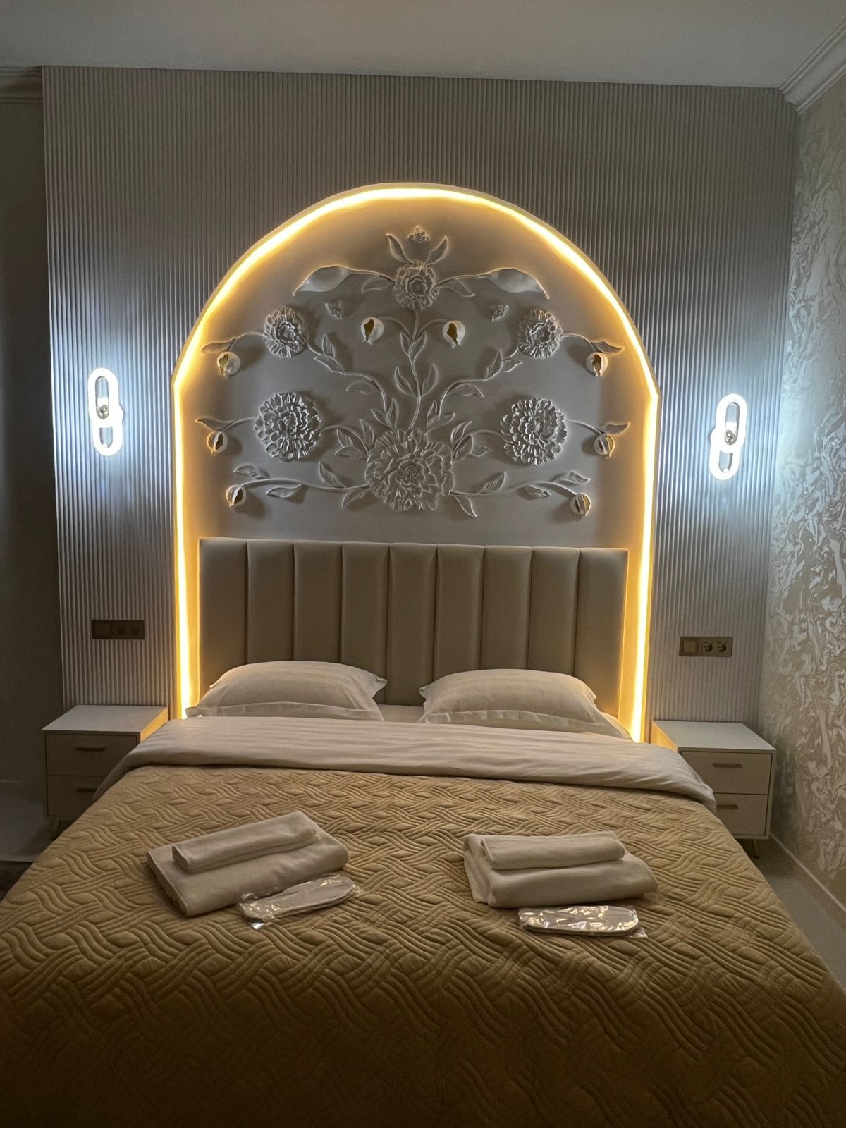 A bedroom with a large bed, a decorative headboard, bedside tables, and a wall with vertical lines.