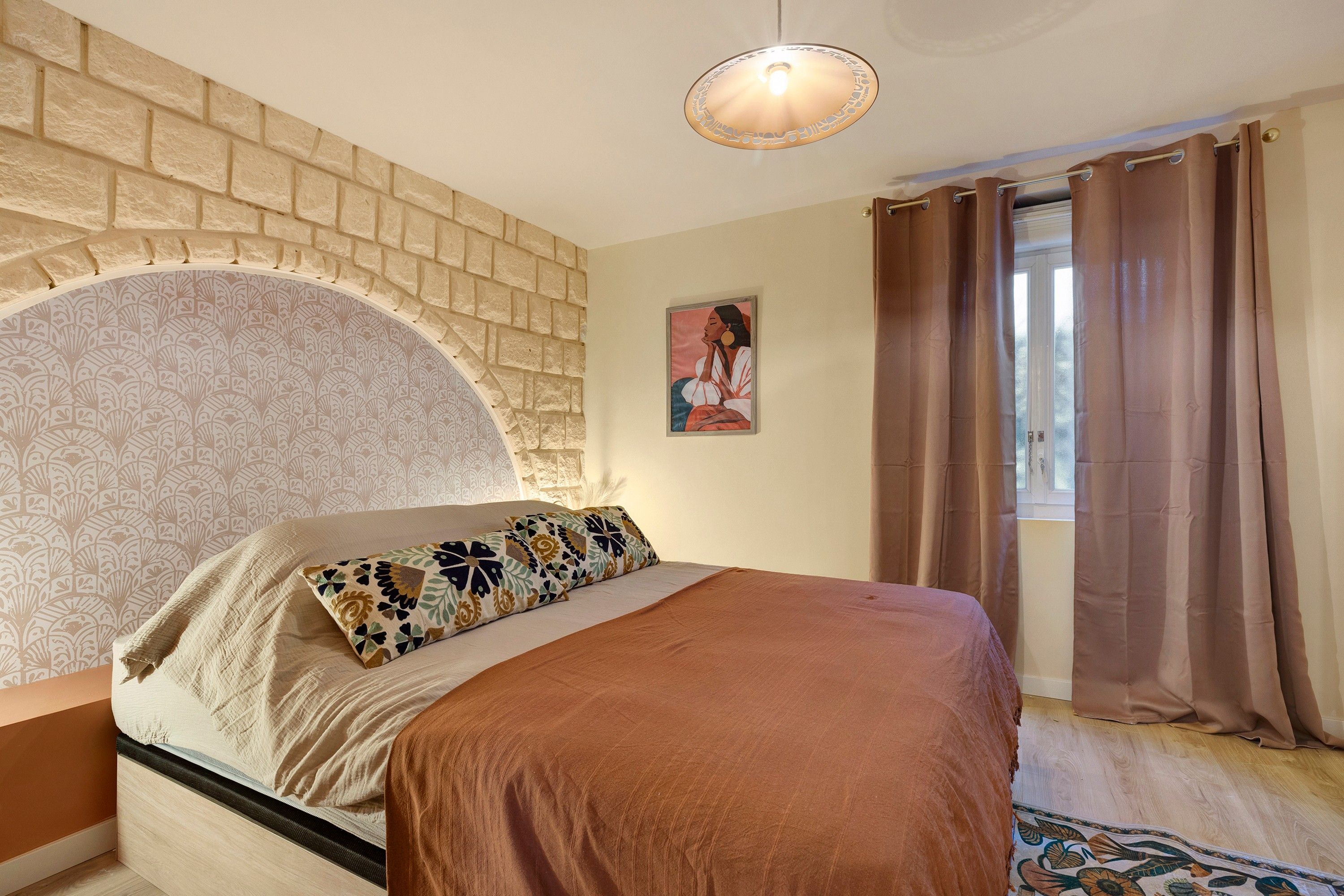 A bedroom with a brick wall, a bed with a patterned pillow, a painting on the wall, and a window with curtains.