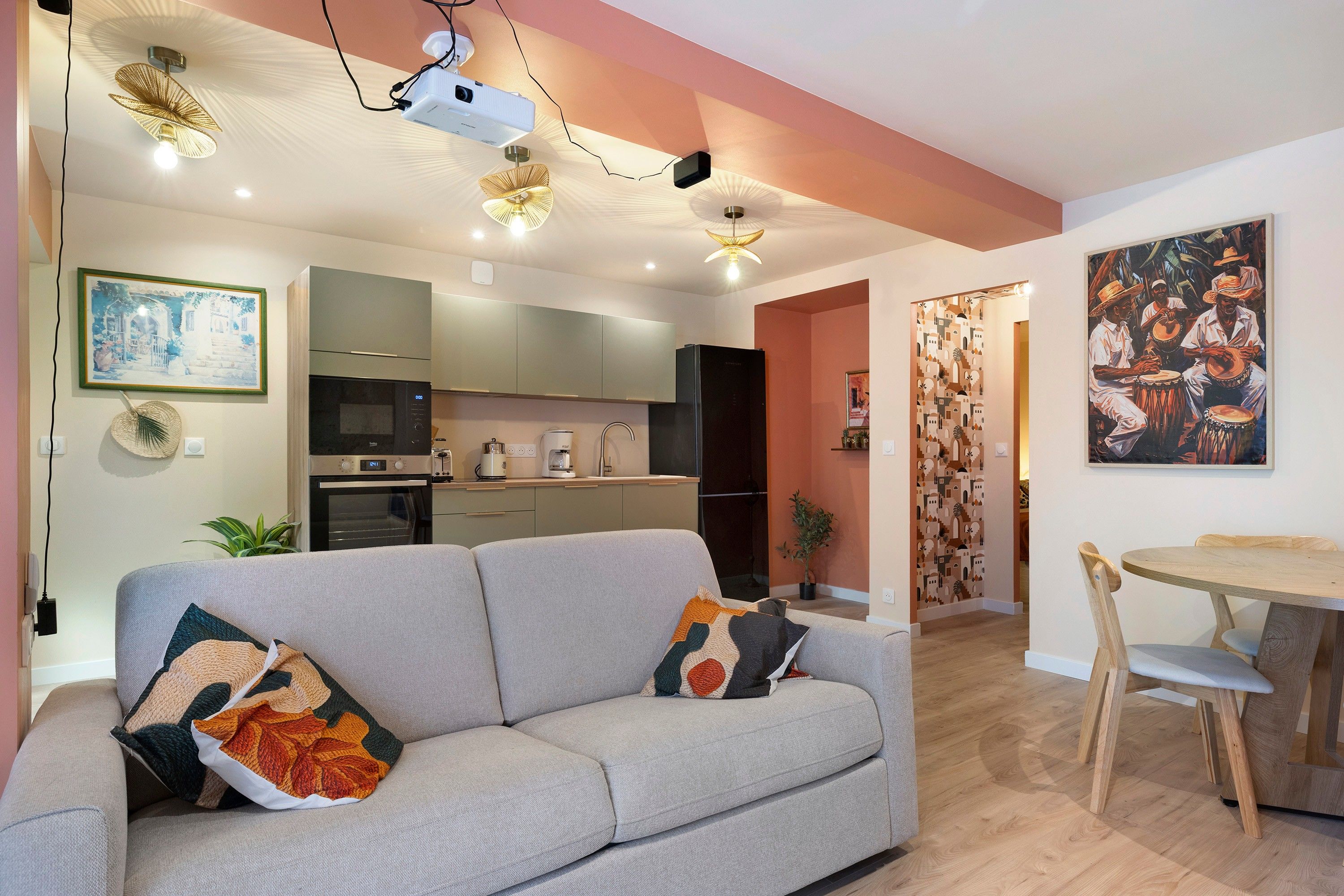 Een moderne woonkamer met een grijze bank, een eettafel met stoelen, een keukenhoek en een plafondprojector.