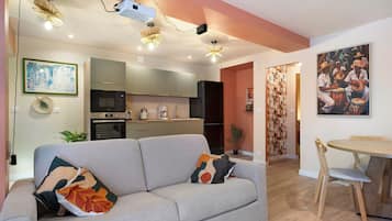 Een moderne woonkamer met een grijze bank, een eettafel met stoelen, een keukenhoek en een plafondprojector.