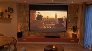 A living room with a large flat-screen TV mounted on a stone wall, a wooden dining table, and a small potted plant. - Casa Calma – Private Spa & Cinema (Bordeaux)