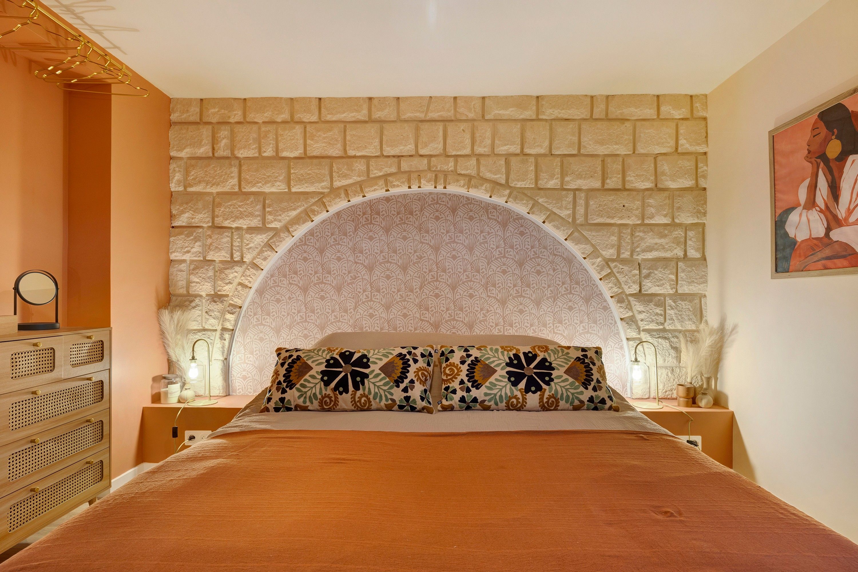 A bedroom with a stone wall arch, a bed with patterned pillows, a dresser, and a wall-mounted lamp.