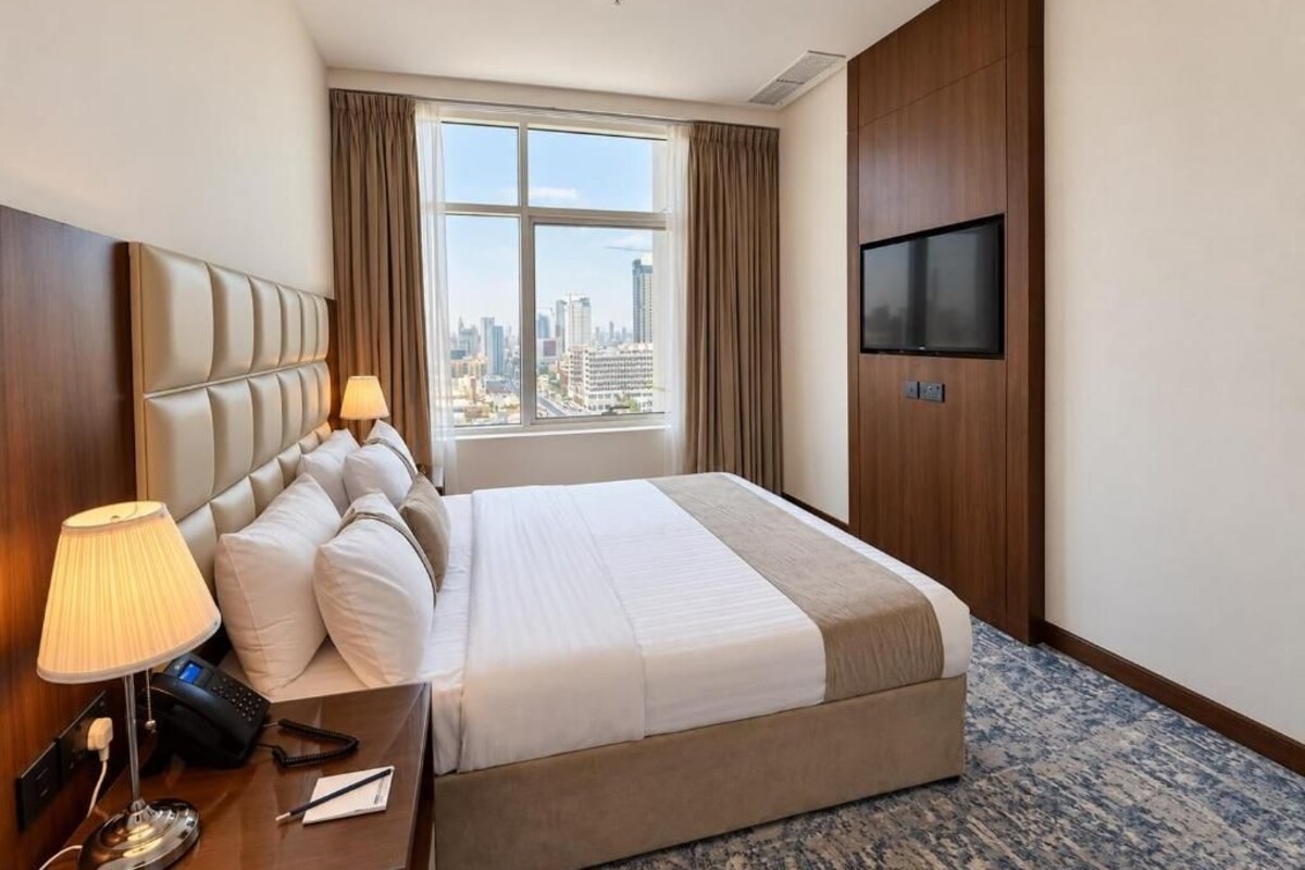 A hotel room with a bed, a TV, a desk with a lamp, and a view of a cityscape through a large window.