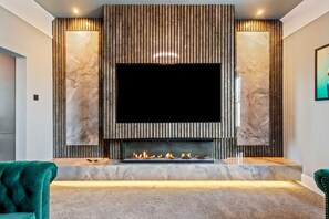 Ein modernes Wohnzimmer mit einem großen Flachbildfernseher, der über einem Kamin angebracht ist und von Marmorplatten flankiert wird, mit einem bequemen grünen Sessel.