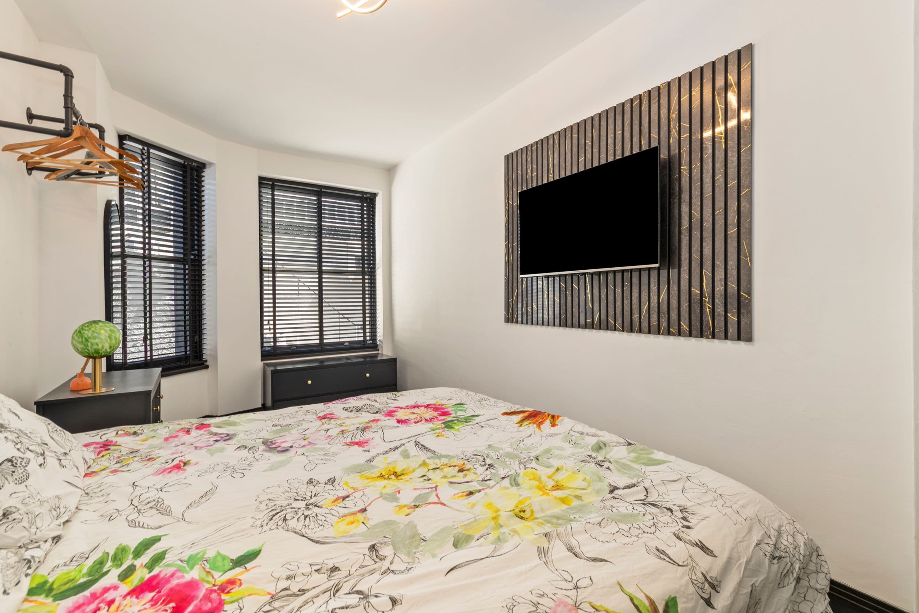 Quarto com cama, duas janelas com persianas, TV fixada na parede e luminária pendente.