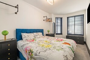 Un dormitori amb un panell de fons blau, un edredó amb estampats florals, una làmpada verda rodona i un prestatge negre.