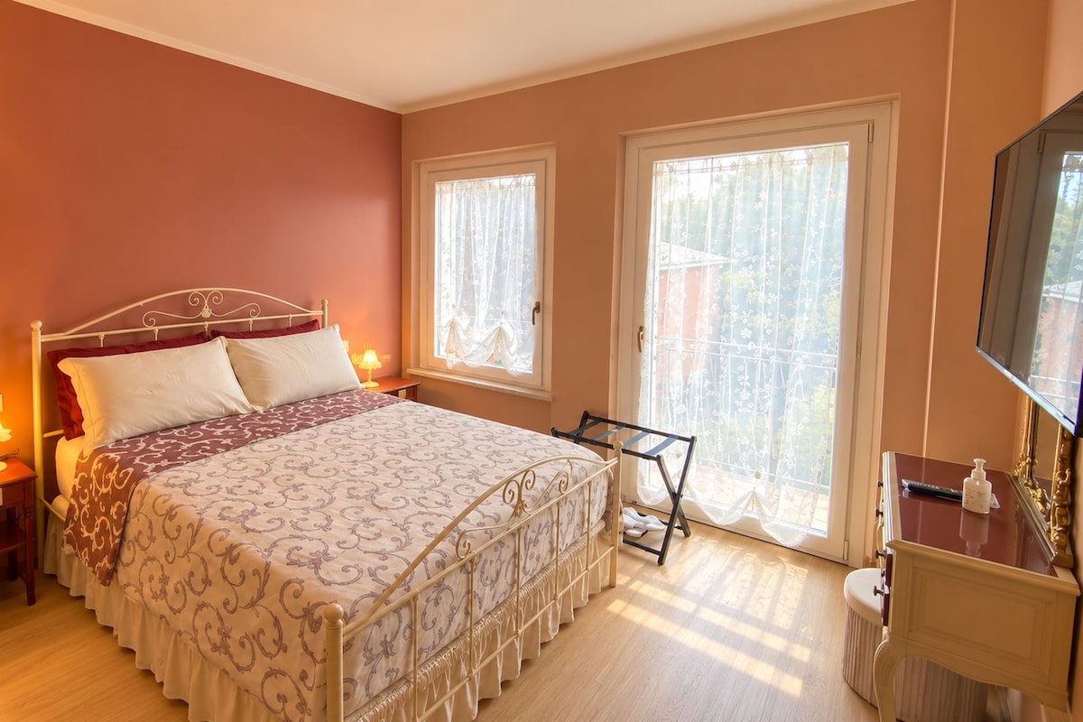 A bedroom with a four-poster bed, a bedside table with a lamp, a dresser, and a large window with curtains.