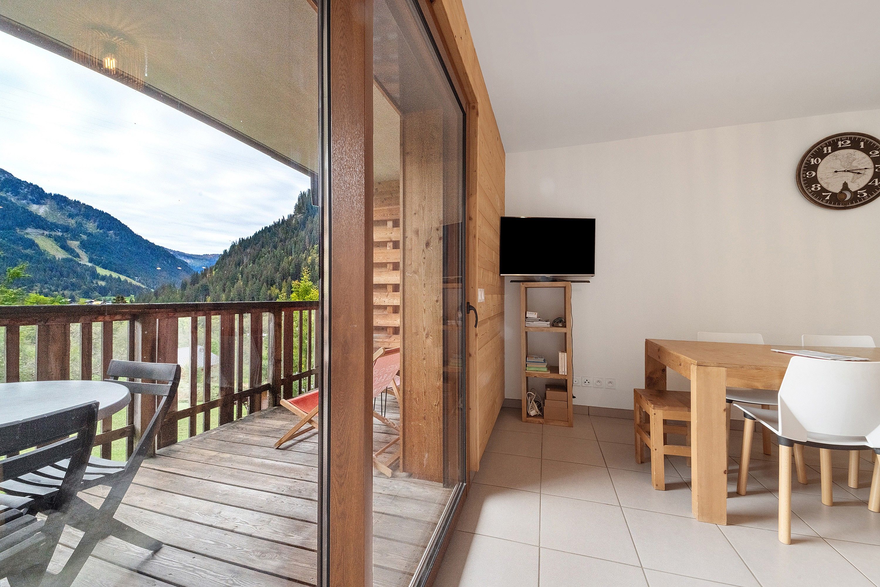 Ein Balkon mit Holztisch und -stühlen, einem an der Wand befestigten Fernseher und Blick auf Berge.