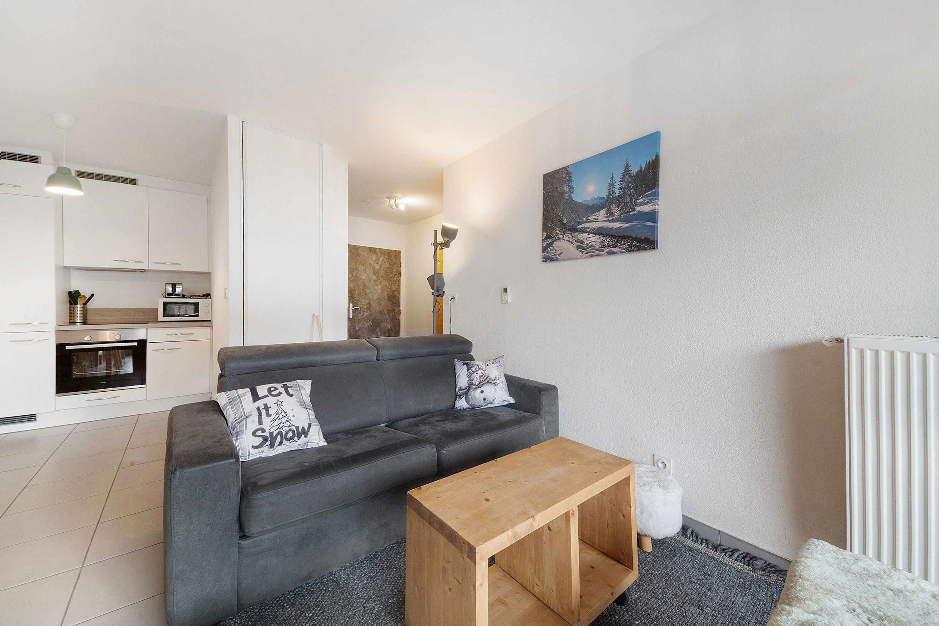 Ein modernes Wohnzimmer mit grauem Sofa, einem Holztisch und einer Küchenzeile mit weißen Schränken und Geräten.
