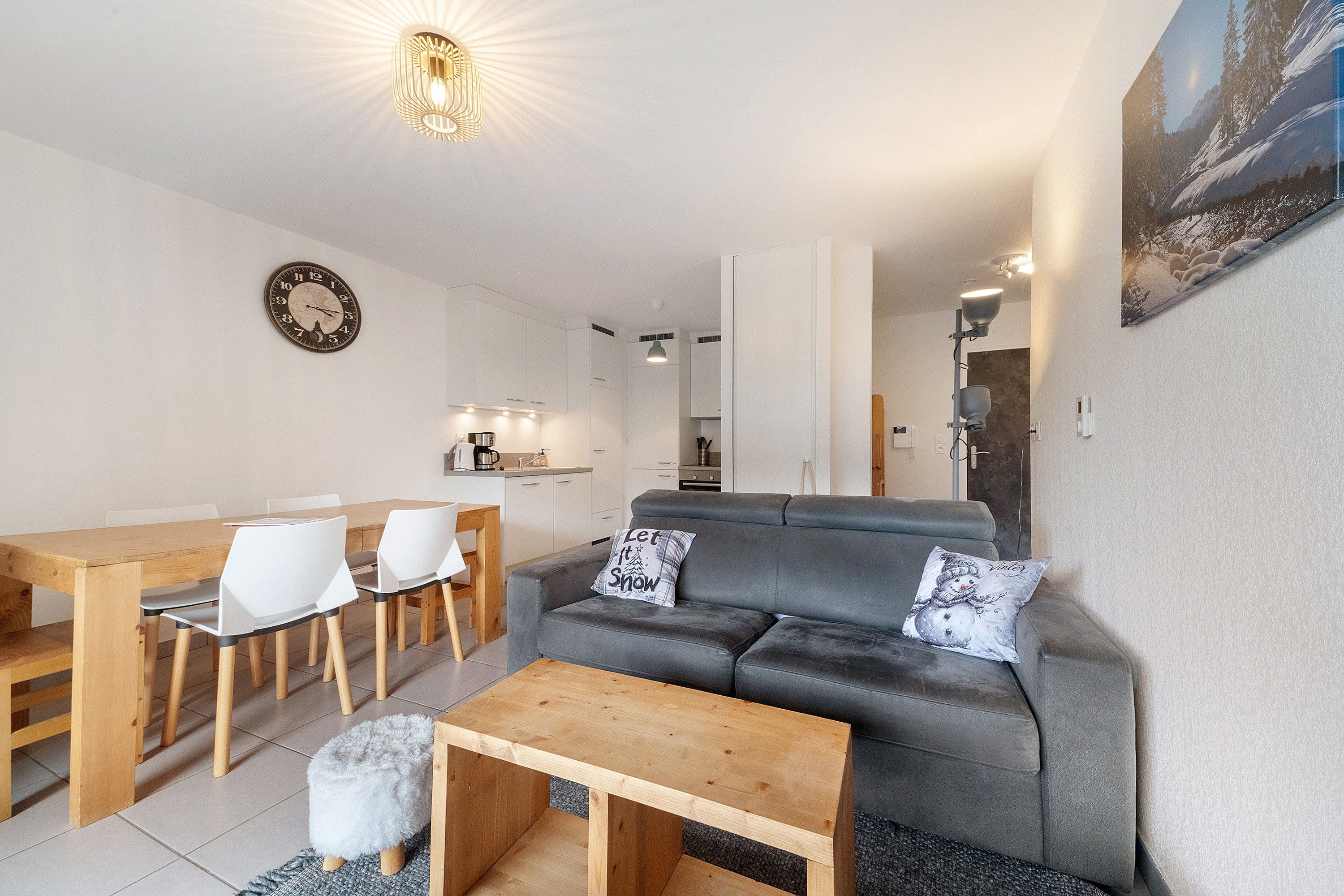 Ein modernes Wohnzimmer mit einem grauen Sofa, einem Holztisch und weißen Stühlen. Im Hintergrund ist eine Küche mit weißen Schränken und einer Kaffeemaschine erkennbar.
