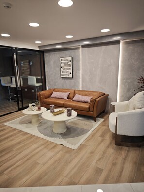 Una sala moderna con un sofá de cuero marrón, dos sillones blancos, una mesa de centro y un separador de vidrio.